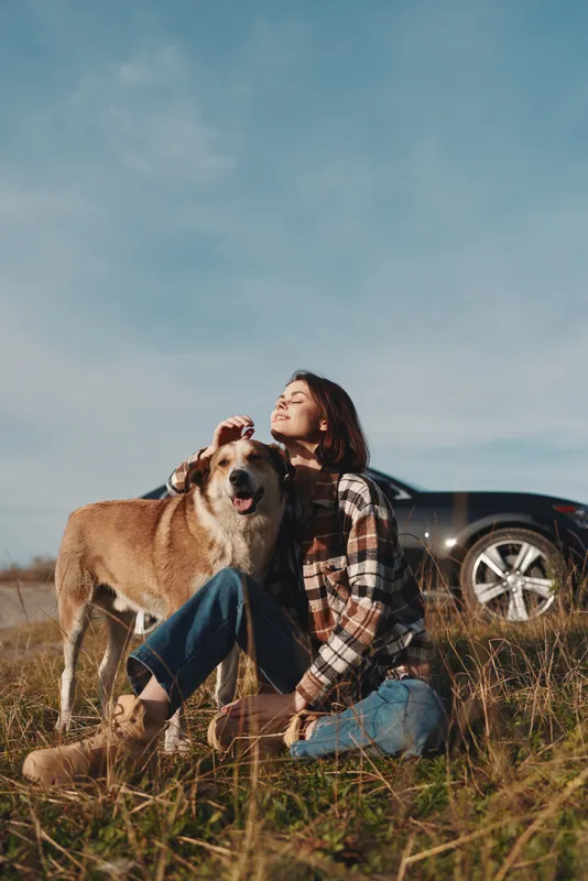 Woman sitting with her dog in a sunlit field beside a parked car, enjoying a peaceful outdoor moment in casual plaid and jeans, facial expressions relaxed, breeze warm, rural vibe and companionship.