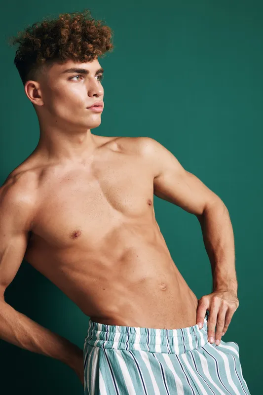 Young man with curly hair posing shirtless in striped pants against green background, showing athletic build and confident expression in studio portrait.