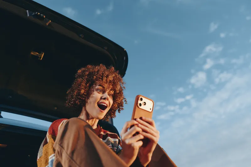 Cheerful woman with curly hair taking a selfie with a phone near a car on a sunny day, outdoor lifestyle scene. Natural light and candid smile convey freedom and joy.