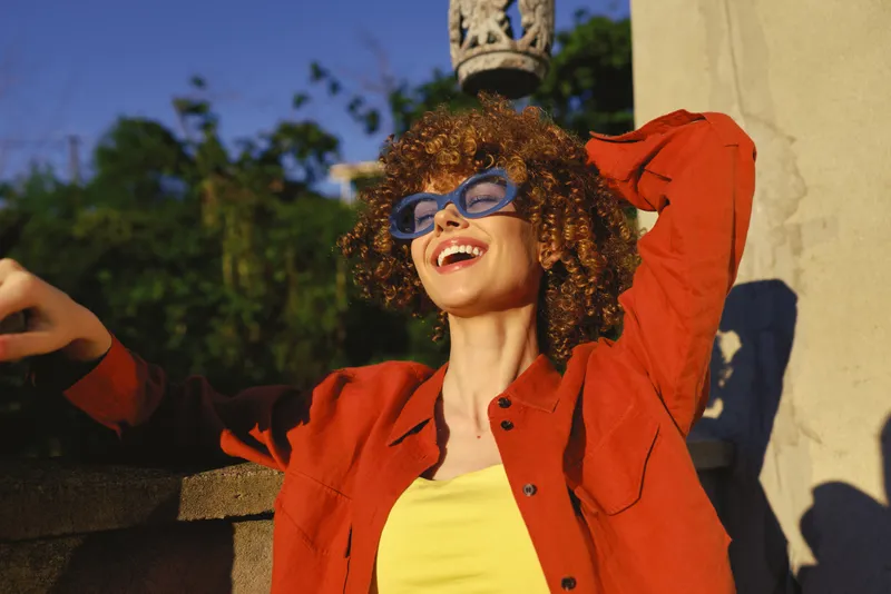 Young woman with curly hair wears a red jacket and yellow top, arms raised in a sunny outdoor setting, radiating carefree joy and confidence
