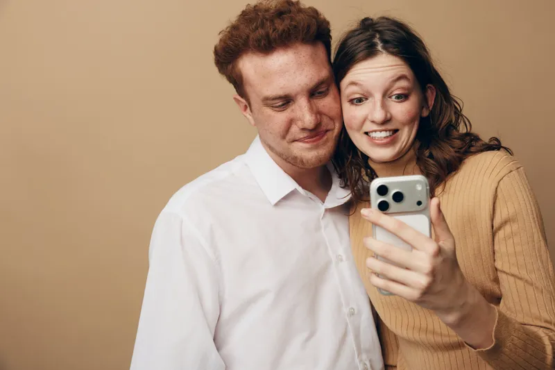 Happy young couple looking at smartphone screen together, smiling and enjoying moment. Casual clothing and neutral background create warm, intimate atmosphere of shared experience.