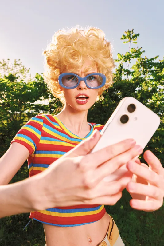 Young woman with retro curls takes a carefree selfie outdoors in a striped crop top and blue sunglasses, capturing a playful sunny moment with a bold pose.