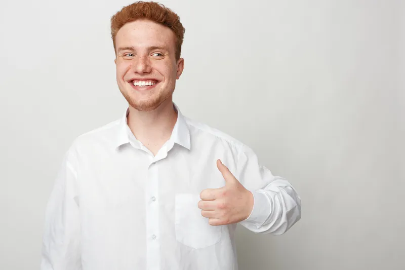Smiling young man with red hair and beard wearing white shirt shows thumbs up gesture. Positive emotion, approval, and confidence concept on light grey background. Friendly casual portrait.
