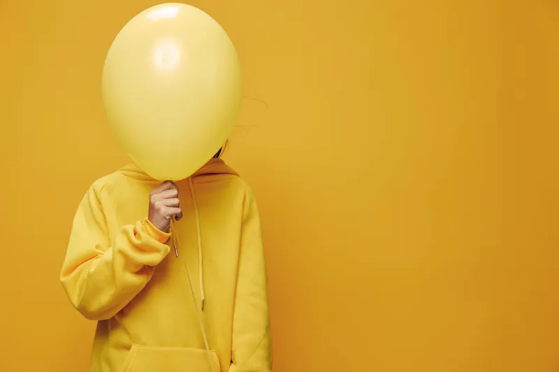 Person in yellow hoodie holding yellow balloon in front of face standing against solid yellow background for creative fun and anonymity concept.