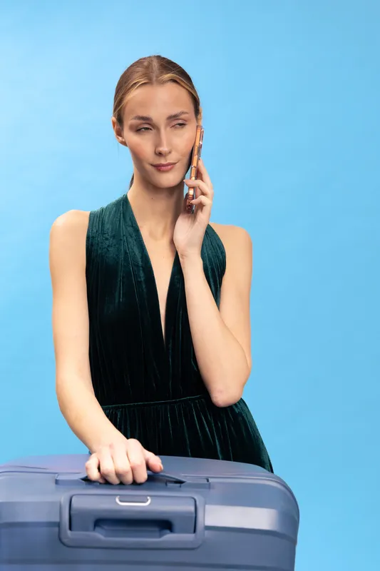 Young woman in green dress holding suitcase and talking on phone. Isolated on blue background. Concept of travel, communication, and planning a trip or vacation.