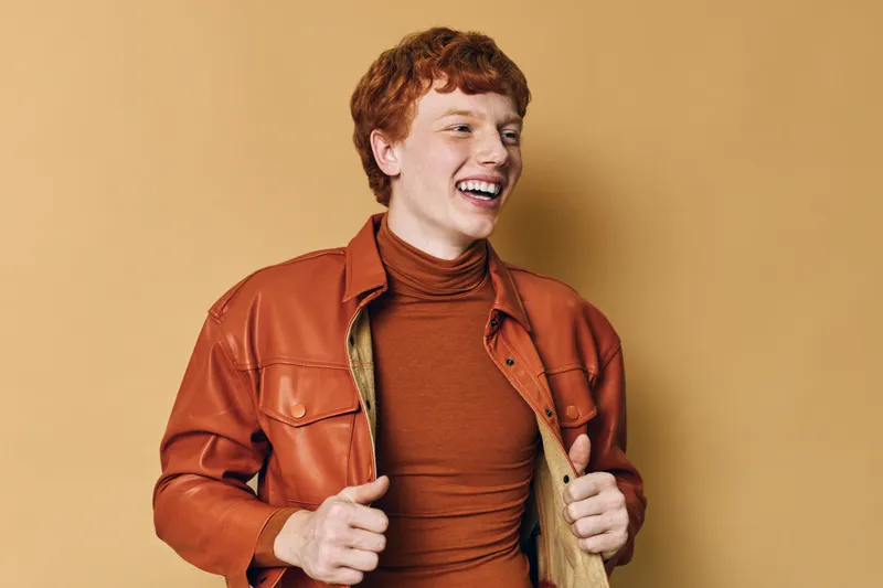 Young man with red hair and freckles wearing a brown leather jacket and turtleneck smiles happily on beige background in studio. Portrait, fashion and style concept.