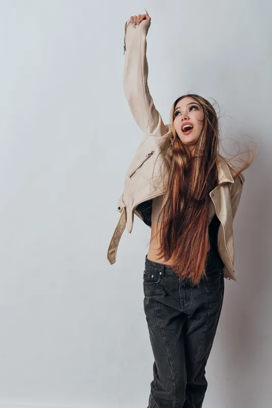 Young woman with long hair wearing leather jacket and black pants raises her arm excitedly against gray background in casual modern fashion style portrait.