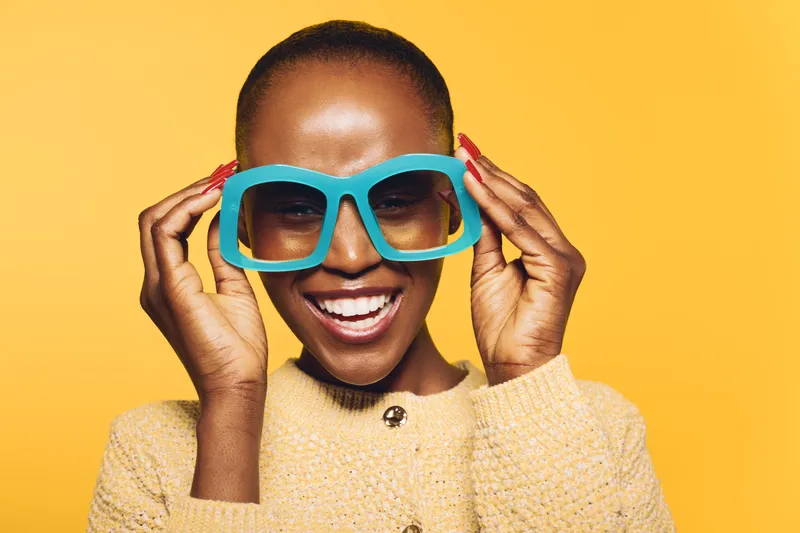 Happy African woman with dark skin wearing oversized blue glasses and beige sweater smiles joyfully on yellow background. Concept of fashion, style, happiness, and beauty.