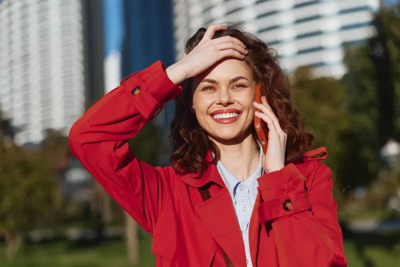 Young woman with curly hair smiles joyfully while talking on a smartphone outdoors in a city park on a sunny day wearing a red coat and white shirt.