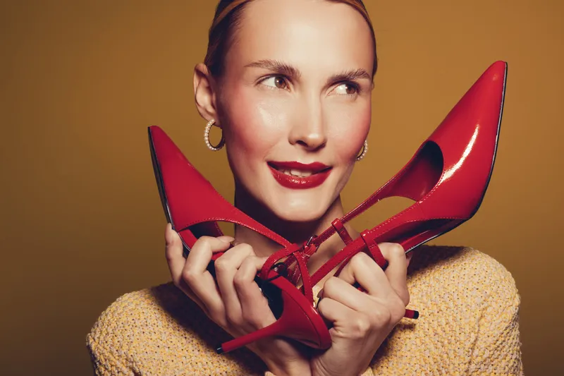 Young woman with red lipstick holds red high heel shoes near face, smiling and looking aside. Fashion concept with bright makeup and stylish accessories on warm background.