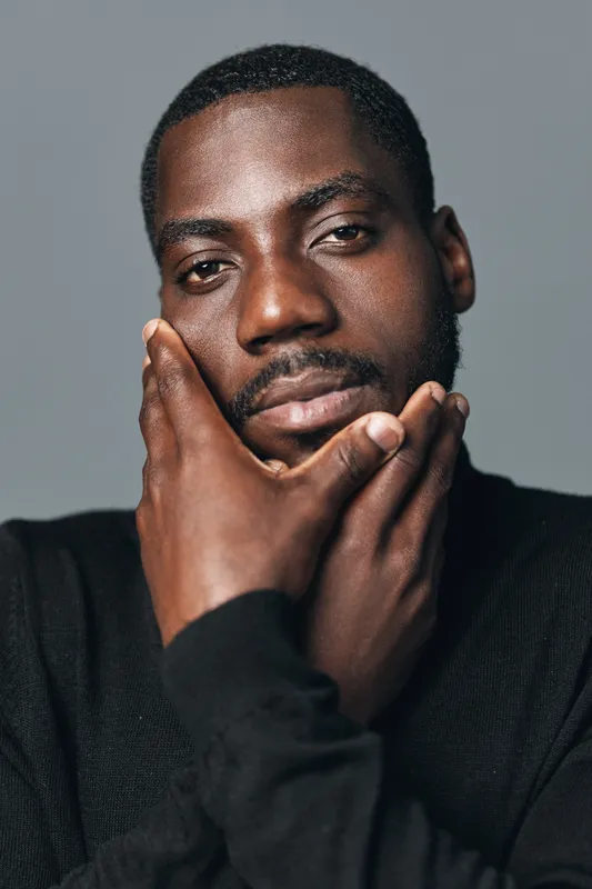 African man with dark skin and beard in black sweater posing with hand on face, creative conceptual portrait with visual style and thoughtful expression on gray background.