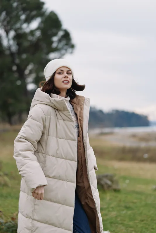 Woman in a long beige puffer coat and white beanie stands in an open field with trees, enjoying cool weather and a relaxed outdoor fashion moment, wind in hair and calm expression.