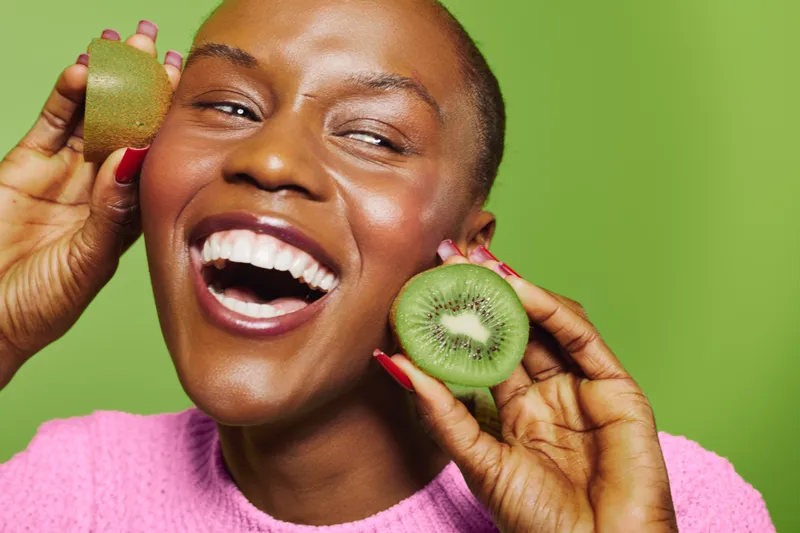 Happy black woman with dark skin laughs holding two halves of kiwi fruit near face. Bright green background contrasts with pink sweater, creating fresh and joyful mood. Concept of healthy lifestyle.
