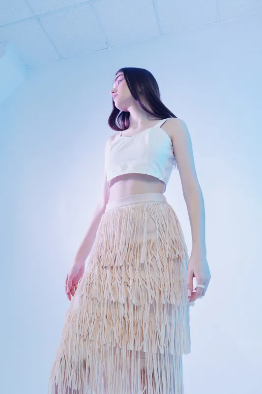 Young woman in a white crop top and layered fringed skirt poses against a soft blue background. The fashion portrait highlights beauty, style, and modern clothing trends.