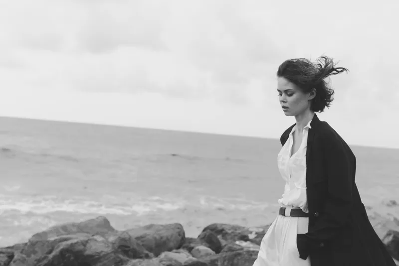 Woman on a windy coastline wearing a white dress and dark coat, thoughtful expression by the sea, monochrome mood, fashion and solitude amid rugged rocks