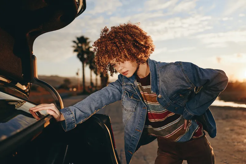 Woman lifestyle outdoors, opening car door at sunset, curly hair and denim jacket, warm golden light, casual street moment, relaxed smile, preparing to travel