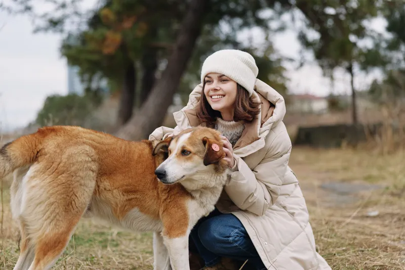 Woman, dog, outdoors in a park, warmth and companionship on a chilly day, wearing a cozy coat and knit hat, smiling beside a loyal canine in nature