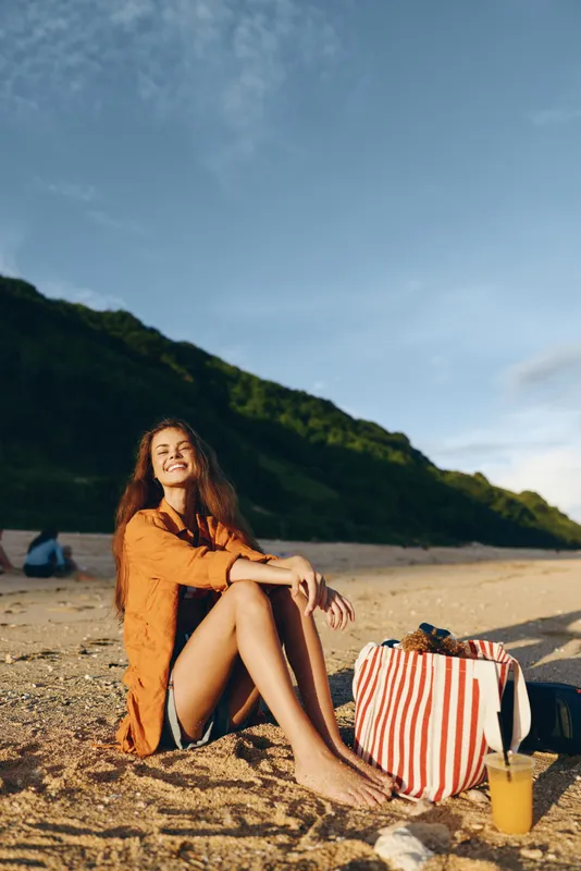 Young woman with long hair wearing an orange shirt sits barefoot on the sandy beach smiling under a clear blue sky with green hills in the background.