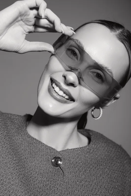 Smiling Caucasian woman with fair skin wearing transparent glasses and hoop earrings. Close-up portrait with stylish outfit and natural makeup on neutral background.