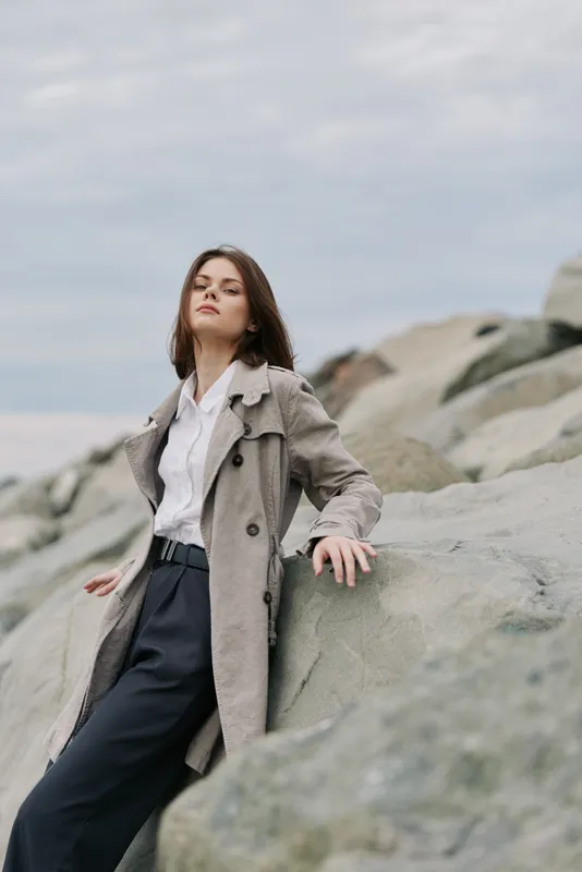 Young woman in beige trench coat and white shirt stands on large rocks under cloudy sky. Casual fashion, outdoor portrait, confident pose, natural light, autumn style concept.