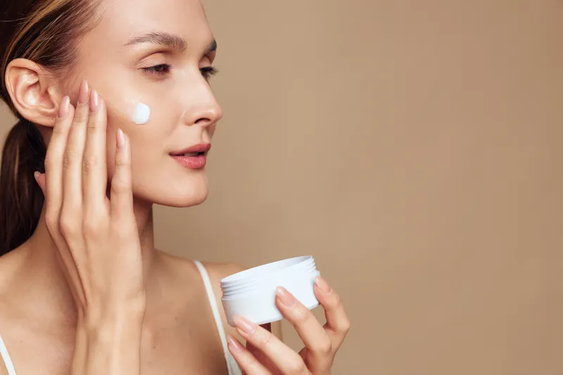 A young woman applies moisturizing cream on her cheek with fingers while holding a white jar against a plain beige background. Concept for skincare and beauty routine.