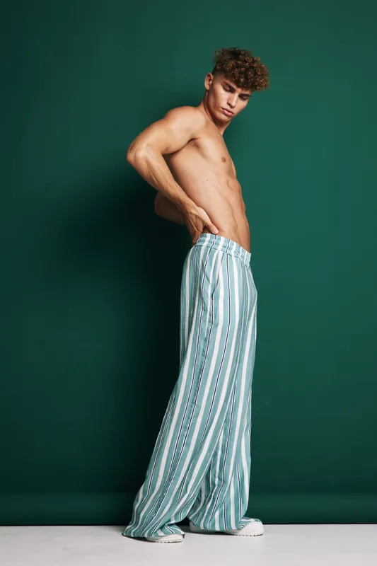 Young man shirtless with curly hair posing in striped loose pants against dark green background along standing casual fashion model. Fitness youth style natural look portrait.