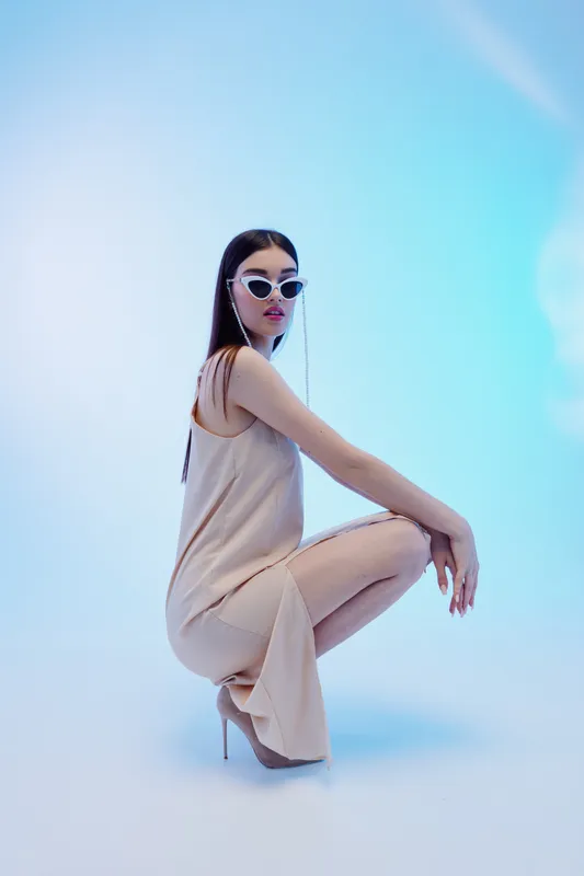 Stylish woman in a beige dress and high heels poses wearing trendy sunglasses. Studio shot on a soft blue background. Concept of woman beauty fashion and elegance.