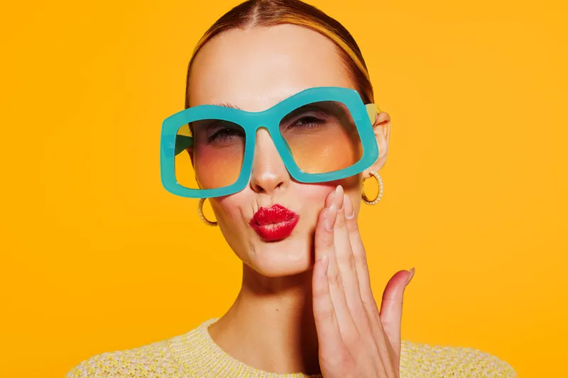 Young woman with large blue sunglasses and red lipstick makes a kiss face on bright yellow background. Fashion portrait with playful expression and colorful style concept.