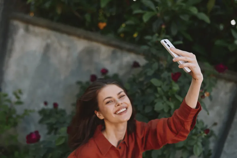 Smiling woman in a red top takes a selfie outdoors in a garden, capturing a lifestyle moment. Bright natural light creates a cheerful, casual mood with warm tones and soft focus