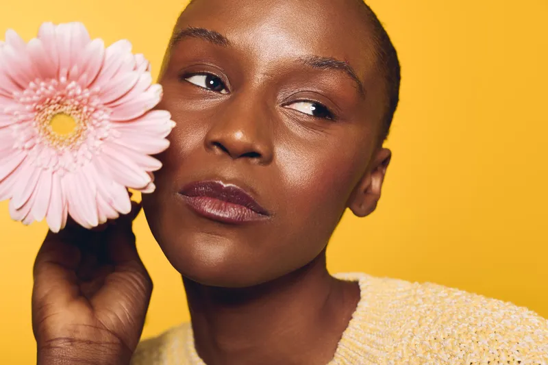 Close-up of a thoughtful black woman with dark skin holding a pink flower near her face against a bright yellow background. Concept of beauty, skincare, and natural wellness.
