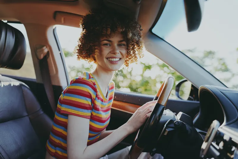 Vibrant woman in a striped shirt captures a lifestyle moment while driving a car, smiling with joy, casual pose, colorful retro vibe, warm sunlight through the window