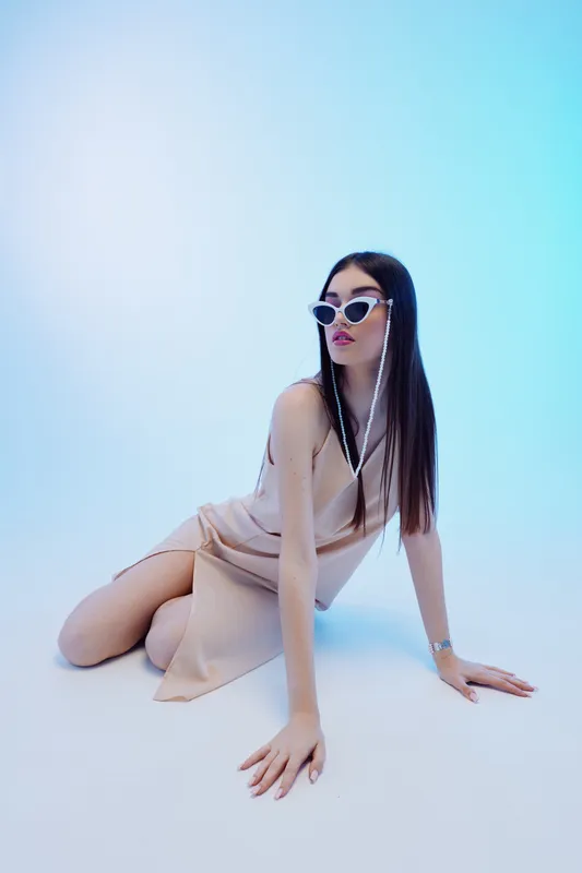 Young woman in a beige dress and sunglasses poses on the floor against a soft blue background. Concept of beauty, fashion, style, and elegance.