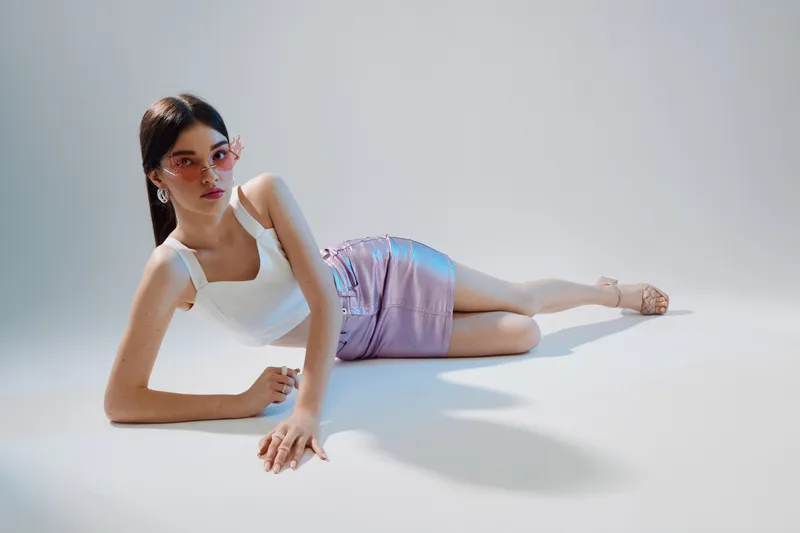 Young woman in white top and purple skirt lies on floor wearing pink sunglasses. Studio shot with soft lighting. Concept of woman beauty fashion and modern style.