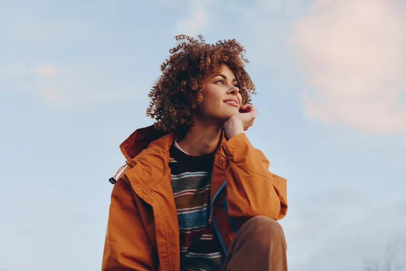 Woman lifestyle outdoors, curly hair, orange jacket, sky background, one hand touching hair, relaxed thoughtful smile, soft daylight, casual fashion, natural colors, calm atmosphere