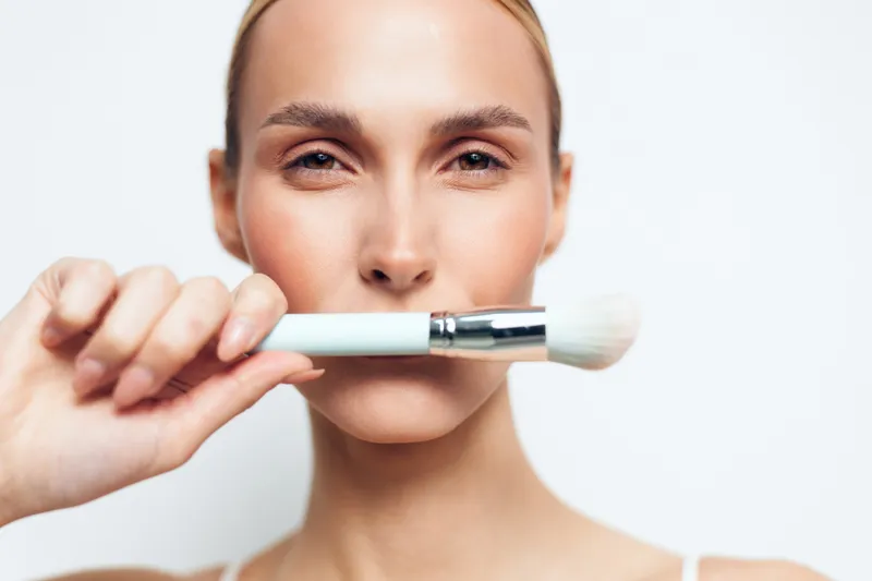Young woman with blonde hair holds a white makeup brush close to her face while looking confidently at the camera against a plain white background.