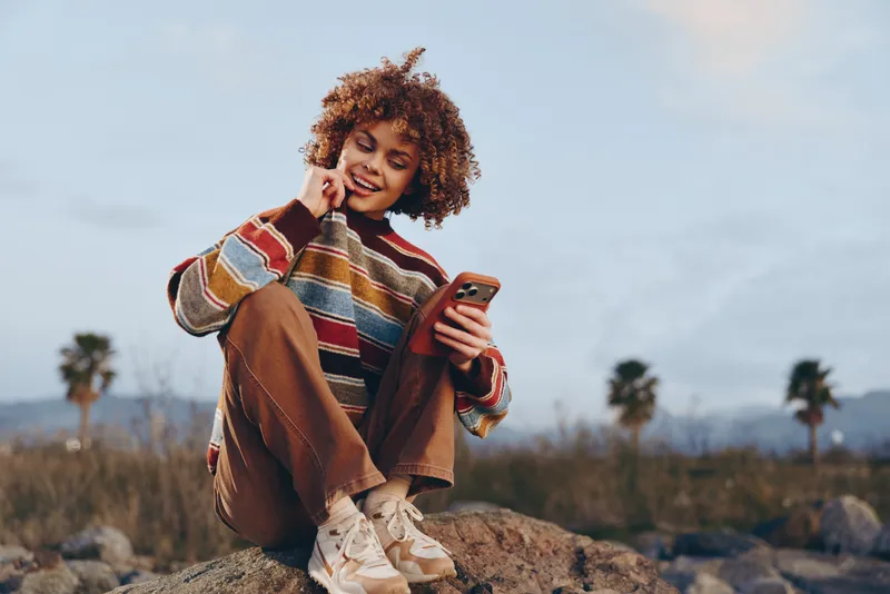 Woman lifestyle smartphone outdoors smile sitting rock casual nature sky, young woman laughs holding phone on rocks, relaxed travel moment under cloudy horizon, natural background