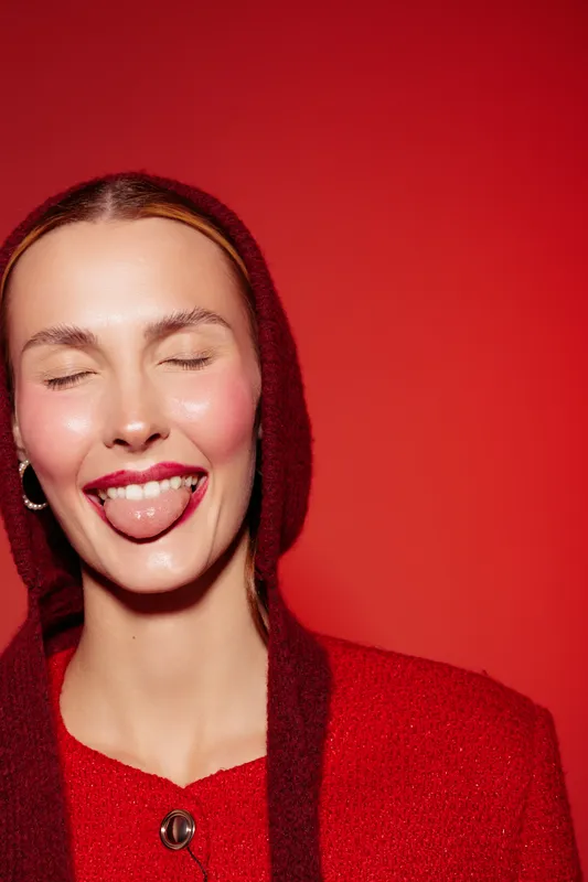 Young woman with closed eyes and tongue out wears red hooded sweater against red background. Playful expression and bright makeup create fun and vibrant mood.