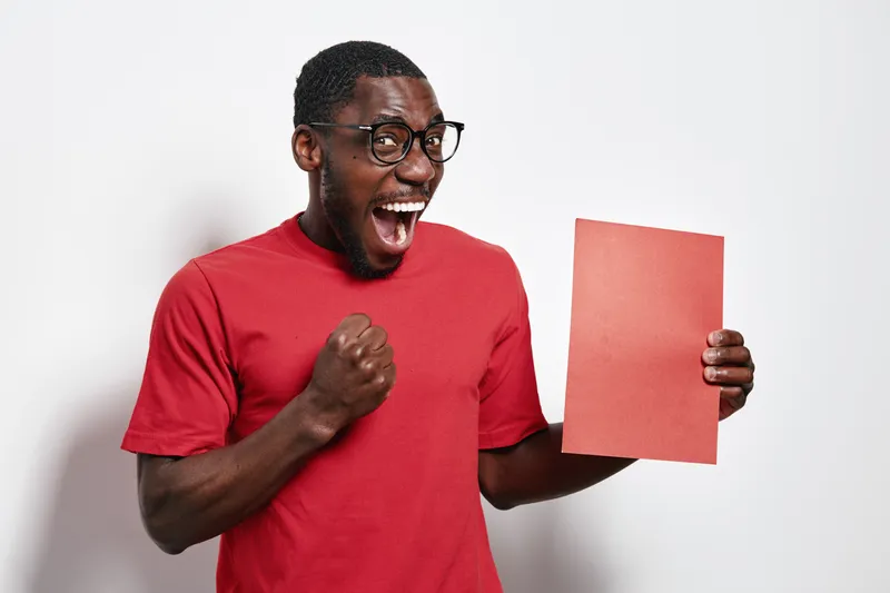 African American man with dark skin wearing glasses and red shirt holding red paper with excitement and joy on white background. Happy black male expressing success and celebration.