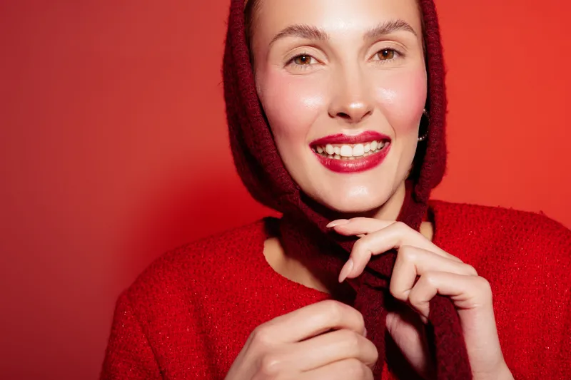 Smiling woman in red sweater and scarf poses against red background. Close-up portrait with bright lipstick and natural skin glow. Concept of fashion, beauty, and winter style.