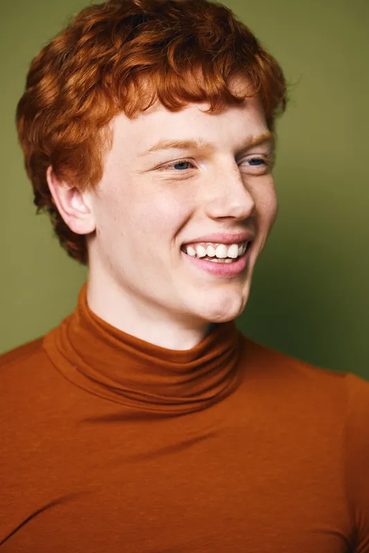 Young man with red hair and freckles smiling dressed in a brown turtleneck against a plain green background. Portrait showing happiness and natural expression with casual clothing.