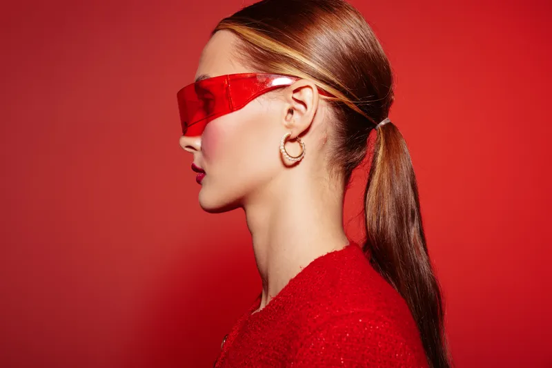 Profile of a young woman with red sunglasses and ponytail against red background. Fashion portrait with bold colors and modern accessories in studio lighting.