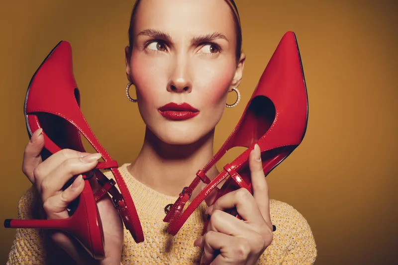 Woman with red lipstick holds red high heel shoes near face, looking sideways. Fashion concept with bold makeup and stylish accessories on warm brown background.