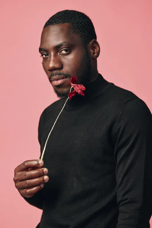 African man with dark skin holding red flower near face in creative conceptual visual style. Portrait with pink background and black turtleneck sweater for artistic expression and emotion.