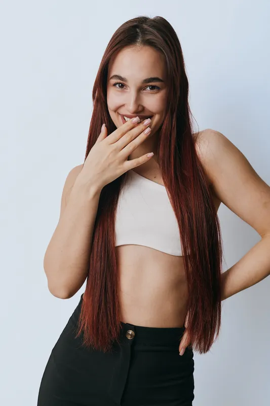 Young woman with long straight hair smiling and covering mouth with hand wearing white crop top and black pants on light background. Portrait, casual, happy and confident.