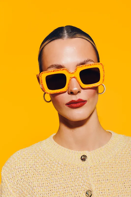 Stylish woman with slicked hair wears oversized orange sunglasses and hoop earrings against bright yellow background. Fashion portrait with bold colors and confident expression.