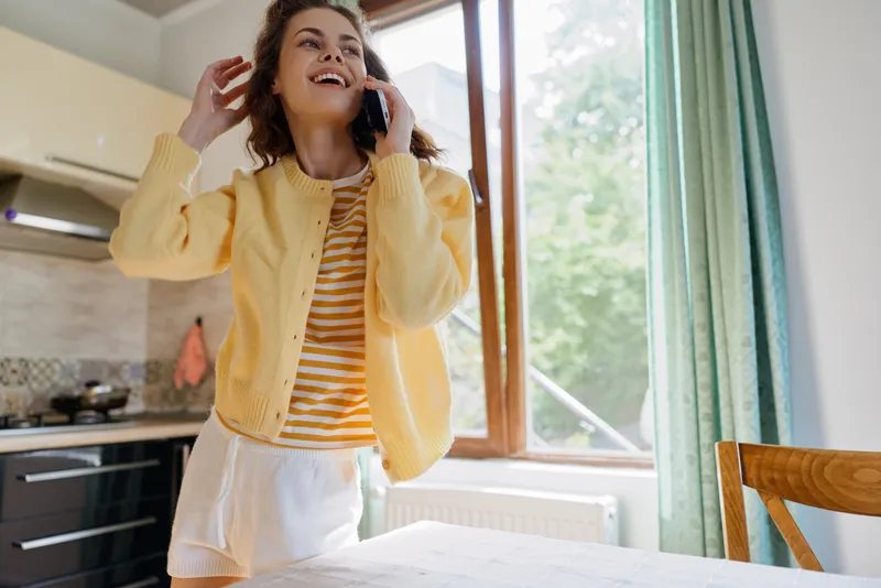 A cheerful woman in a yellow cardigan stands in a bright kitchen, talking on a smartphone, mid conversation. A casual home lifestyle moment, daytime sunlight streaming through a window.
