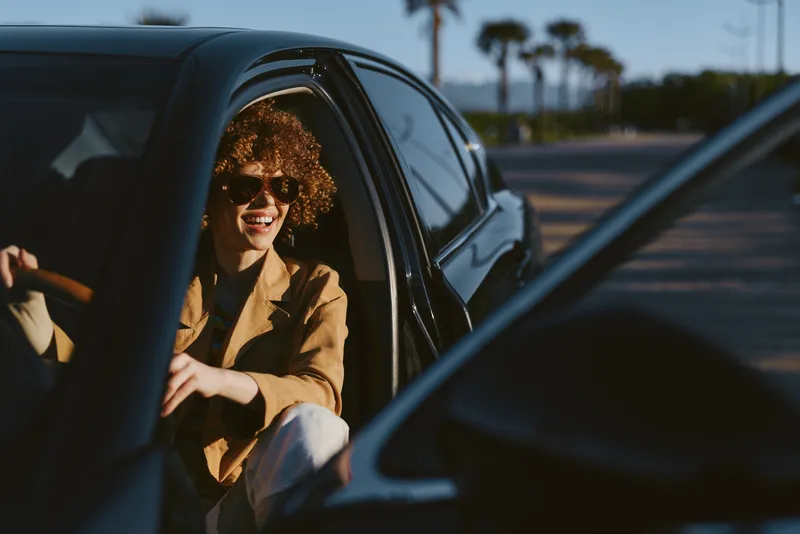 Smiling woman with curly hair sits in a car wearing a brown jacket. Retro film color with warm lighting adds a casual lifestyle mood and road travel vibe.
