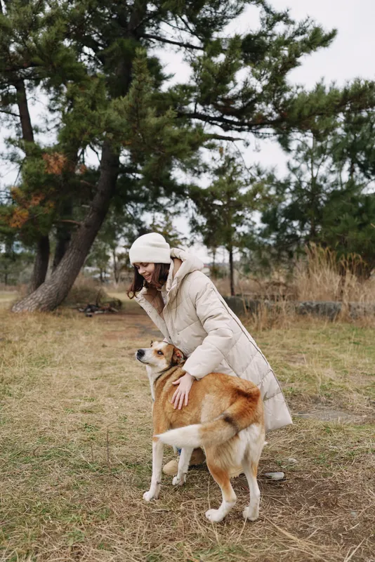 Woman with dog in an outdoor park, dog interaction on a chilly day, wearing a warm coat and beanie, surrounded by trees and grass for a calm natural moment
