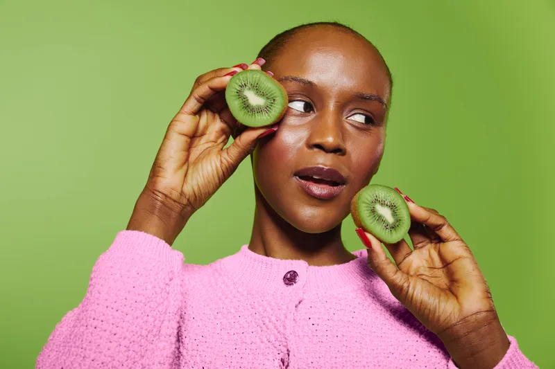 Young African woman with dark skin holds two kiwi halves near her face against green background. Concept for healthy lifestyle, natural food, and fresh fruit nutrition.
