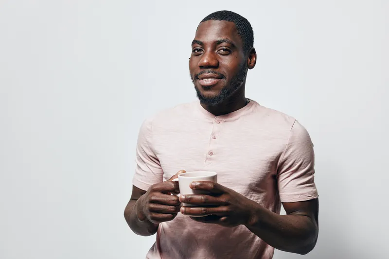 African American man with dark skin holding a cup and smiling, wearing a light pink shirt against a plain white background. Portrait of happy black male with beard.